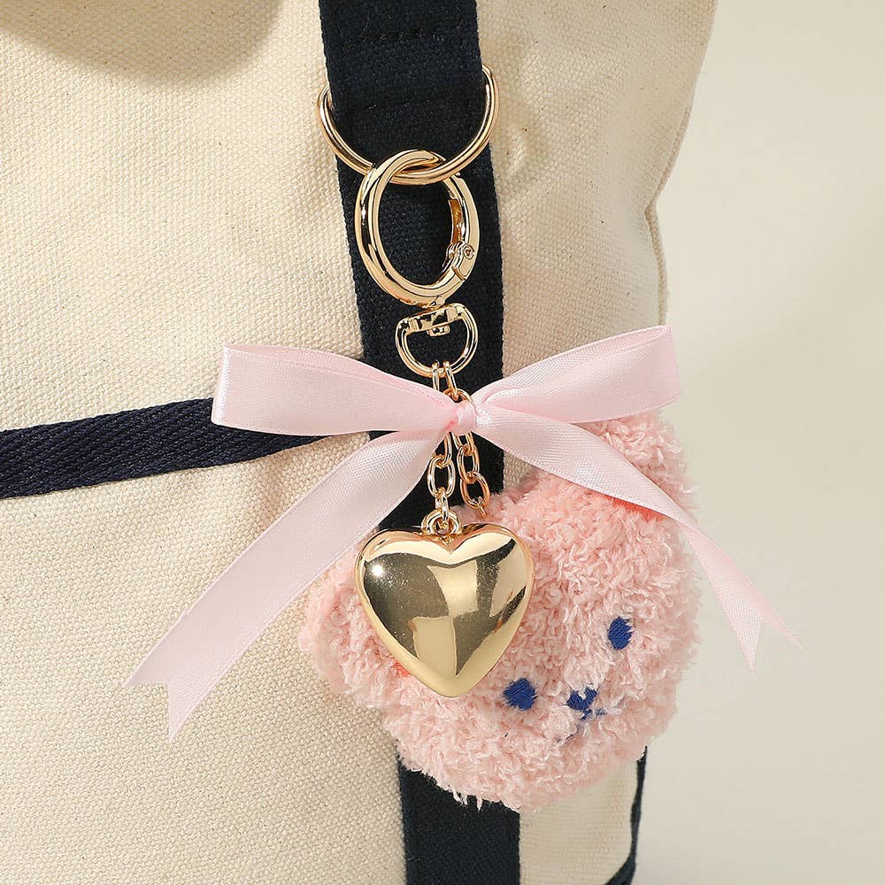 Pink plush bear keychain with a gold heart charm and satin ribbon bow attached to a cream and navy tote bag.